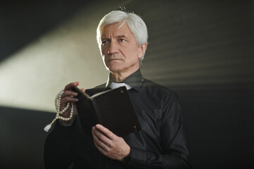 Dramatic portrait of white haired senior priest holding Bible and praying in dark with god rays,...