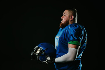 An Asian man with a red beard in a blue american football uniform holds a helmet on a black...