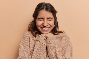 Fototapeta premium Overjoyed Iranian millennial girl with long dark hair keeps hands under chin giggles positively keeps eyes closed dressed in oversized knitted jumper isolated over brown background. Happy emotions