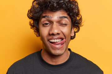 People positive emotions concept. Studio close up of young cheerful amused funny smiling Hindu guy dressed in black casual tshirt standing in centre isolated on yellow background showing tongue