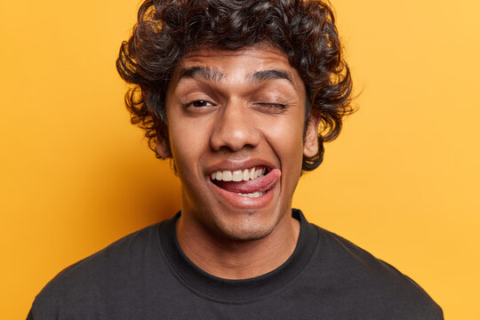 People positive emotions concept. Studio close up of young cheerful amused funny smiling Hindu guy dressed in black casual tshirt standing in centre isolated on yellow background showing tongue