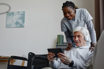 Portrait of white haired senior man with disability relaxing at home and using tablet with nurse...