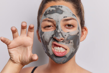 Close up shot of serious European woman clenches teeth and makes paw with hand frowns face applies nourishing clay mask for skin treatment isolated over white background. Beauty procedures concept