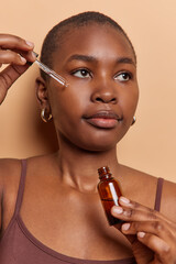 Vertical shot of dark skinned African woman with short hair applies collagen lifting serum puts on...
