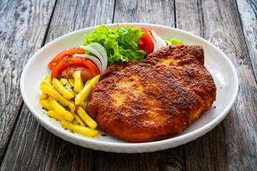 Breaded seared pork chop with fresh vegetable salad and French fries on wooden table
