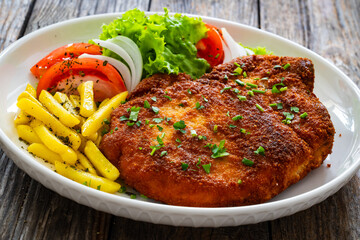 Breaded seared pork chop with fresh vegetable salad and French fries on wooden table
