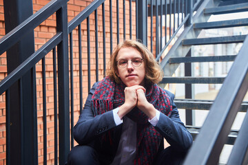 Fototapeta premium A handsome young man in a pantsuit and scarf. European. A college or university student. Street photo set. Sitting on the stairs