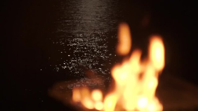 A barbecue grill burns on a campfire in nature at night. Change of focus to the distant plan, to the moonlit path to the sea, the beach. Dark water, waves. Beautiful flames, sparks, smoke. Handheld