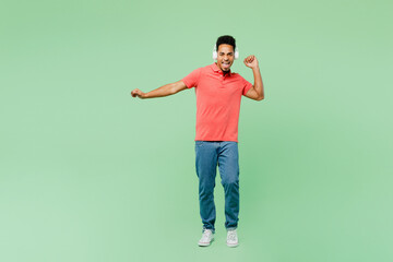 Fototapeta na wymiar Full body fun young man of African American ethnicity he wear pink t-shirt listen to music in headphones dance raise up hands isolated on plain pastel light green background studio. Lifestyle concept.