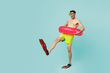 Full body sideways young man wear green shorts swimsuit relax near hotel pool hold in hand inflatable rubber ring flippers isolated on plain blue background. Summer vacation sea rest sun tan concept.