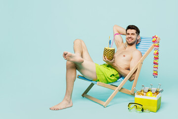 Full body young happy sexy man wear green shorts swimsuit relax near hotel pool sit in deckchair drink pineapple cocktail isolated on plain blue background. Summer vacation sea rest sun tan concept.