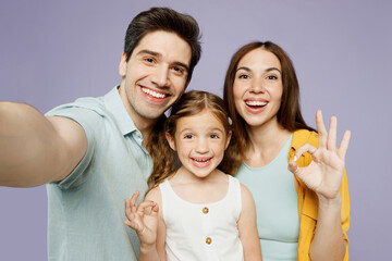 Close up young fun happy parents mom dad with child kid daughter girl 6 year old wear blue yellow casual clothes do selfie shot pov mobile cell phone show ok okay isolated on plain purple background.