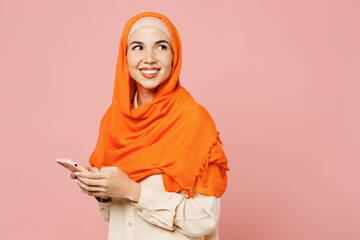 Side view young arabian asian muslim woman wear orange abaya hijab hold in hand use mobile cell phone look aside isolated on plain pink background. People uae middle eastern islam religious concept.