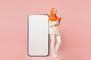 Full body sideways young arabian muslim woman wear orange abaya hijab big huge blank screen area mobile cell phone use smartphone isolated on plain pink background. Uae middle eastern islam concept.