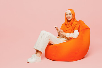 Full body happy fun young arabian asian muslim woman wears orange abaya hijab sit in bag chair hold...