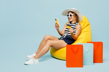 Full body young woman wears casual clothes hat sit in bag chair near shopping package bags use credit card mobile cel phone isolated on plain blue color background. Black Friday sale buy day concept.
