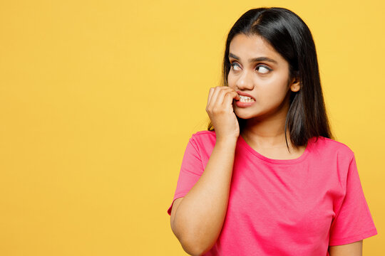 Young minded confused thoughtful happy Indian woman wearing pink t-shirt casual clothes look aside on area biting nails fingers isolated on plain yellow background studio portrait. Lifestyle concept.