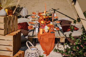 Fototapeta na wymiar Lunch, romantic orange colors. Picnic with blanket, napkin, pillows, candles, wooden decor, dry flowers. Bachelorette party on beach. Boho style design picnic. Decorations for celebration birthday.