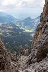 Panoramic view from the Ferrata Passo Santner (Santnerpass-Klettersteig) in the Dolomites, overlooking a deep valley with winding paths, dense forest, and distant mountain ridge under a cloudy sky.
