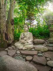 Stone Gautama Buddha decorative statue in a garden under green leaves. Outdoor Garden decor concept. Abakan, vertical view.
