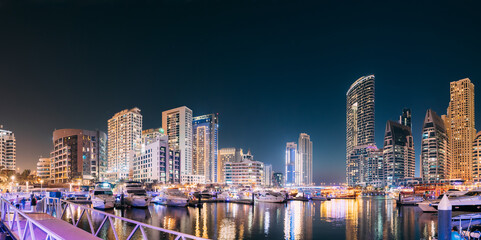 Fototapeta na wymiar Dubai Marina Port, UAE, United Arab Emirates - Beautiful Night View Of Dubai Marina Towers. Yachts Moored Near City Pier, Jetty In Evening Night Illuminations. Panorama, Panoramic View.
