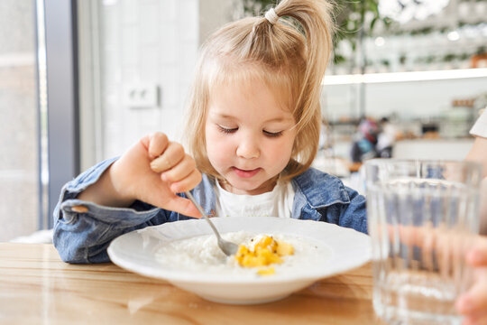 Adorable Girl Drinking Still Water, Eating Rice Porridge With Mango. Enjoying Breakfast. Little Kid Having Breakfast At Cafe. 