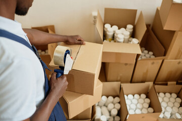Close up of black young man taping boxes while working in packaging department at factory or...