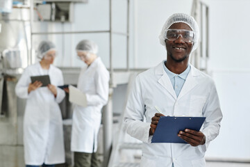 Waist up portrait of black young man wearing lab coat and smiling at camera in clean workshop of...