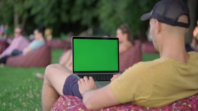 Young Man Using Laptop With Green Mock-up Screen Outside in City Park. Man Watching Videos, Browsing News and Social Networks