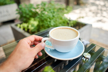 A man drinks coffee, holds a cup of hot Peanut latte in his hand. The table in the coworking space of the coffee shop. A cup of coffee on the veranda on a sunny day. Coffee break at the cafe. Close-up