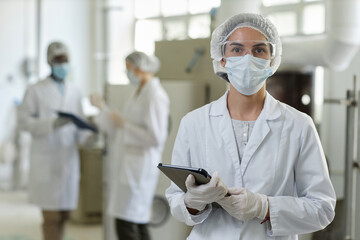Waist up portrait of young woman wearing mask and lab coat looking at camera in factory workshop,...