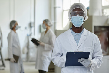 Fototapeta premium Waist up portrait of young black man wearing mask and lab coat looking at camera in factory workshop, copy space