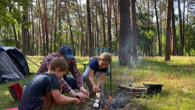 Delightful family camping trip. Father and children gather around campfire to roast marshmallow on wooden sticks in the great outdoors on sunny day. Outdoor cooking while hiking adventure in forest
