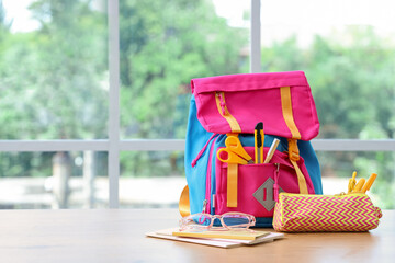 Colorful school backpack with stationery and eyeglasses on wooden table near window in classroom