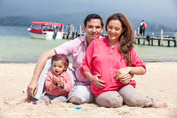 Fototapeta na wymiar Young couple waiting for their second baby having fun with their baby girl at the beautiful white beach of Lake Tota located in the department of Boyaca at 3,015 meters above sea level in Colombia