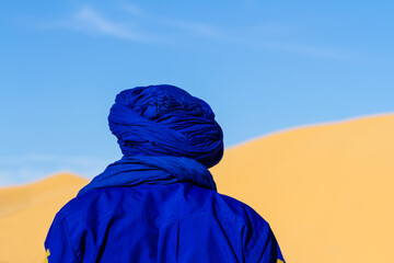 Close-up. Background. Berber in blue traditional clothing against the backdrop of a large dune. View from behind. Tunisia, Africa - obrazy, fototapety, plakaty