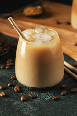 Ice coffee in a tall glass with cream poured over, ice cubes and beans on a dark concrete table.