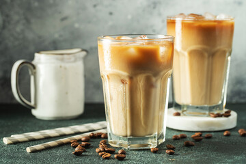 Ice coffee in a tall glass with cream poured over, ice cubes and beans on a dark concrete table.