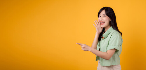 Capture engagement and excitement with Asian young woman wears pastel green shirt on orange shirt show happy smile while placing hand over mouth and pointing with a finger to free copy space.