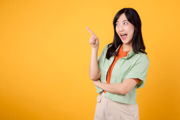 young Asian woman in her 30s, wearing a green shirt on an orange shirt. happy face and pointing finger to free copy space against vibrant yellow backdrop. Unlock savings and shop now!