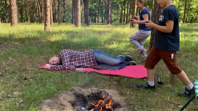 Playful family camping trip in beautiful forest on sunny day. Cheerful kids sneakily paint father face as he naps on mat in the woods. Family bonding time together while vacation in the great outdoors