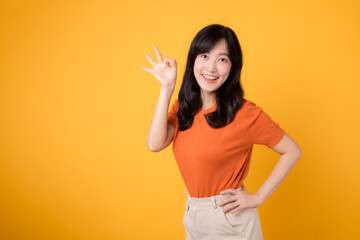 Embodying positivity, young Asian woman 30s flaunts okay sign in orange shirt on yellow background. Hand gesture concept.