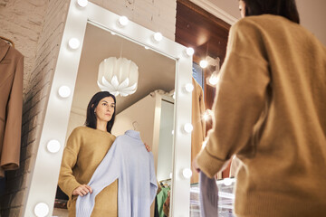 Low angle portrait of adult woman looking in mirror while trying on clothes in boutique, copy space