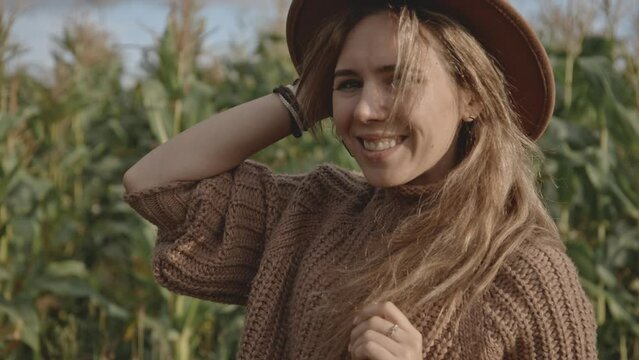 Close up side or profile portrait of blonde hipster smiling and laughing sincerely and broadly while looking into camera. Girl with blonde hair in hat standing in middle of corn field or meadow.