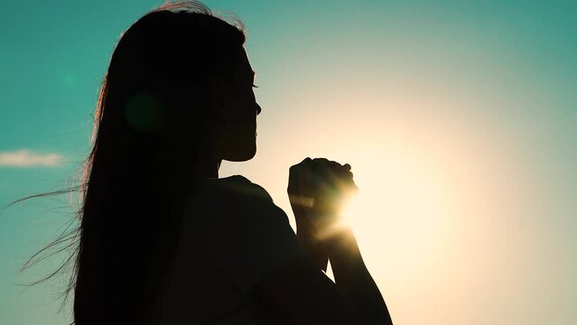 Faith in god. Christian woman praying at sunset close-up. Girl on background of sky in rays of sun prayer to family and children. Relaxation and meditation in nature. Apologize. Hope. Silhouette