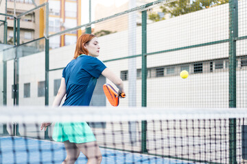 A girl in sportswear is about to backhand a paddle ball during a match on an outdoor court. Concept of women playing paddle. Paddle sport.
