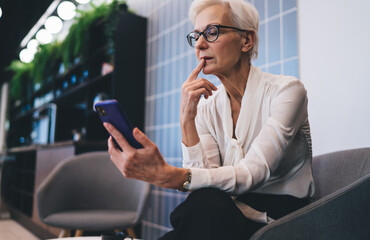 Thoughtful elderly woman using smartphone while sitting in armed chair in office hall