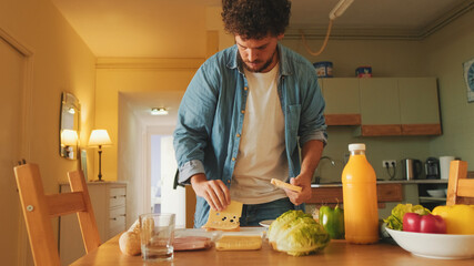 Close-up of the hands of guy dressed in denim shirt cooking sandwich while standing in the kitchen