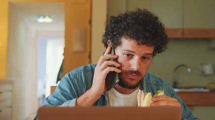 Guy dressed in denim shirt works on laptop while having lunch in cozy kitchen, happily talking on mobile phone