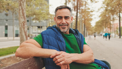 Mature man wearing casual clothes enjoys relaxing on park bench looking at the camera and smiling